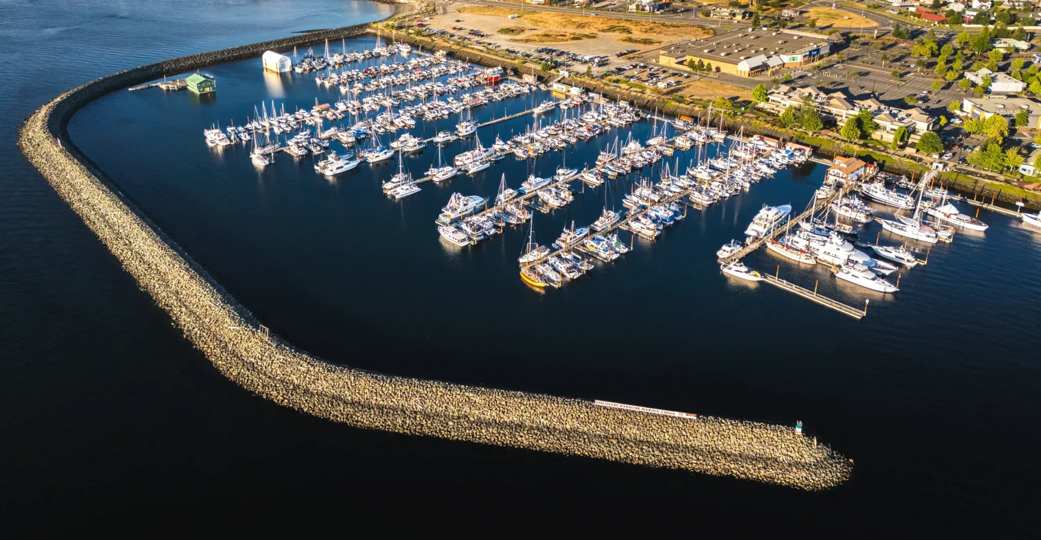 Discovery Marina Harbour in Campbell River,BC. Marina full with boats moored in the ocean, protected by a rock barrier, blue ocean and sunny green landscape.