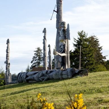 A totem pole stands in a grassy field with yellow flowers in the foreground