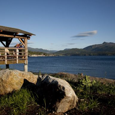 A family stands at the edge of a dock that overhangs a beach and overlooks a large body of water