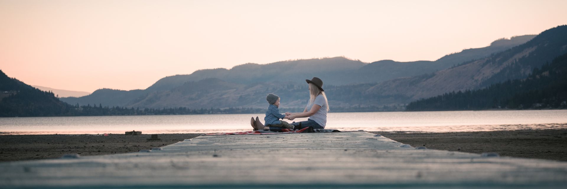 A mother and child sit at the end of a boardwalk and hold hands next to the shore