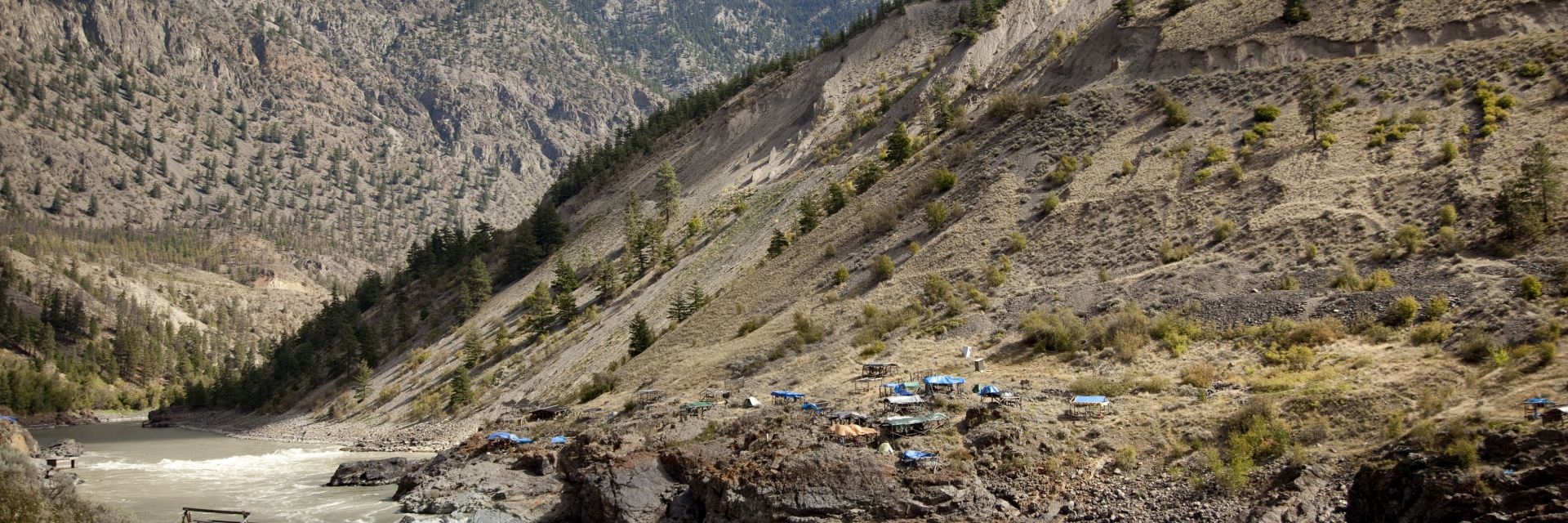 Tents are pitched on both sides of a river at the bottom of hill in Lillooet, BC.