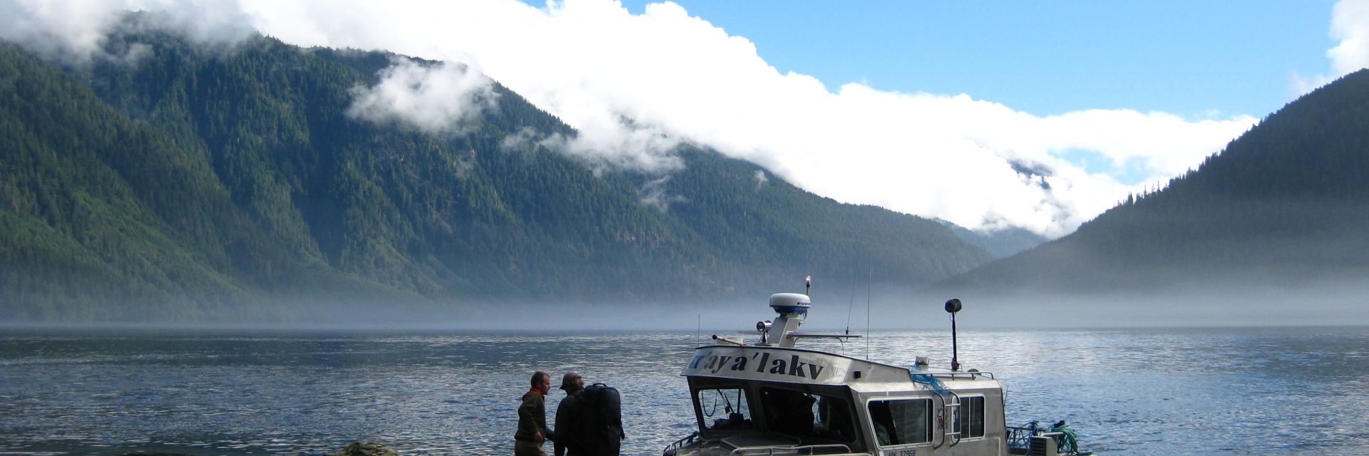 boat docked on water's edge with two people standing on shore and mountains covered in clouds in background