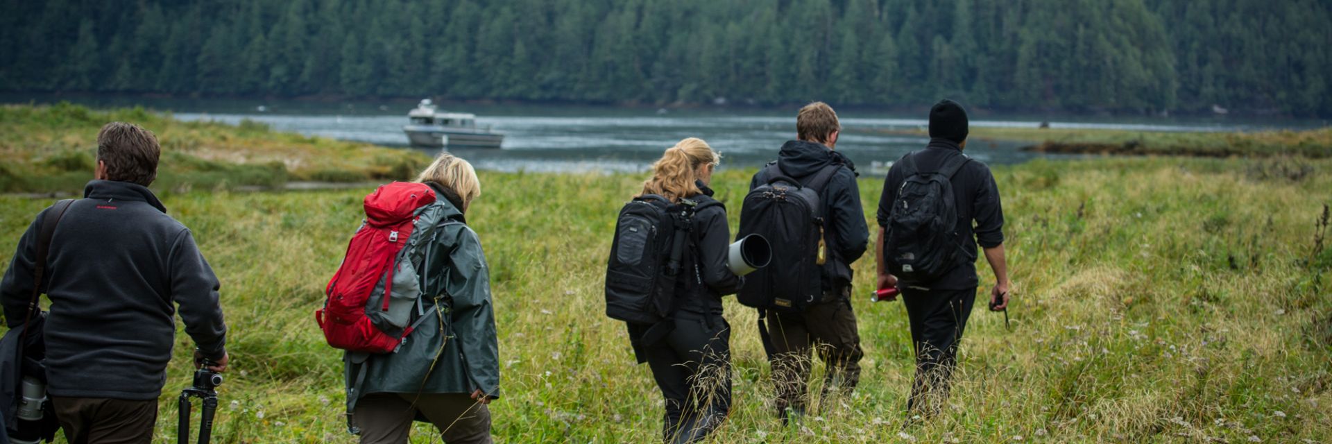 four hikers walk through the tall grass while boat travels in body of water behind them