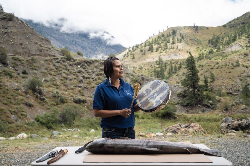 A woman plays a traditional Indigenous drum over the carcass of a fish