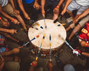 A group of people play a Indigenous drum together in a small circle