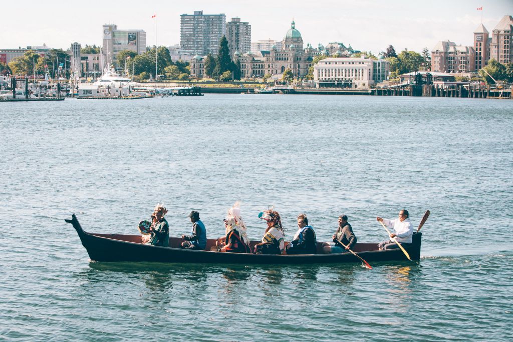 Seven people wearing traditional Indigenous clothing paddle a canoe in the Victoria harbour.