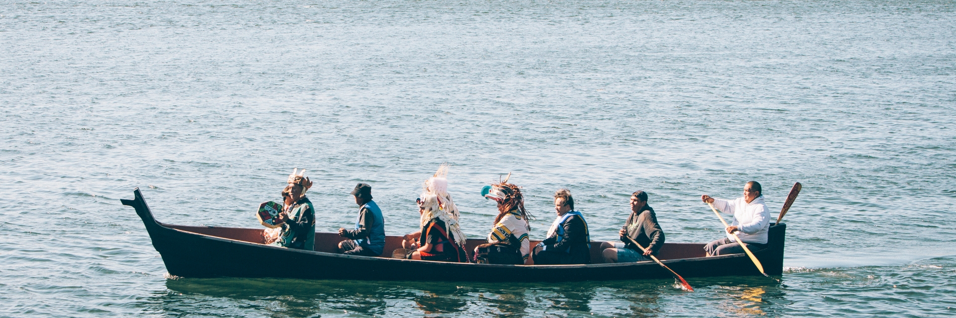 A group of people wearing traditional Indigenous clothing paddle on a canoe in the Victoria harbour.