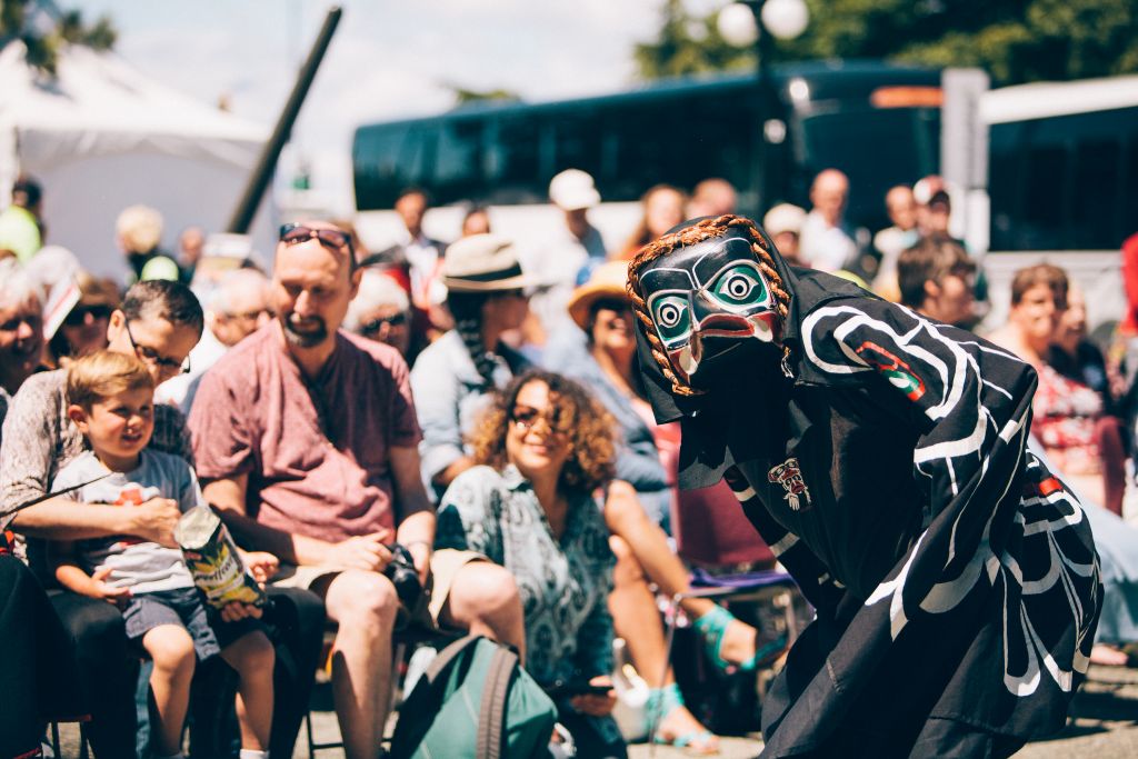 A dancer wearing a mask and traditional Indigenous clothing performs for a crowd