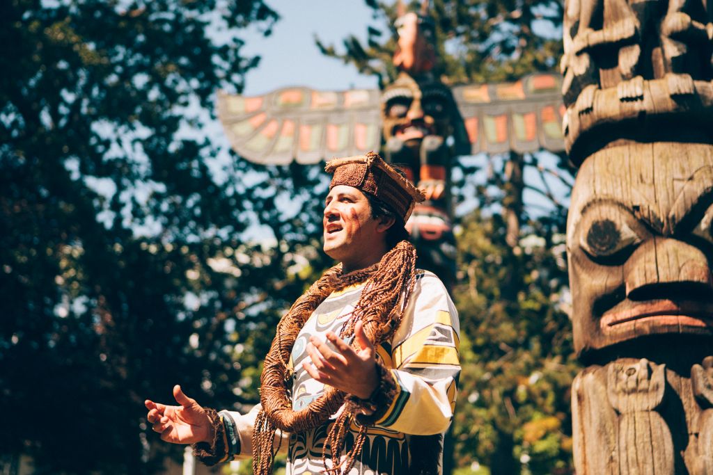 A man wearing traditional Indigenous clothing stands in front of two totem poles while giving a speech