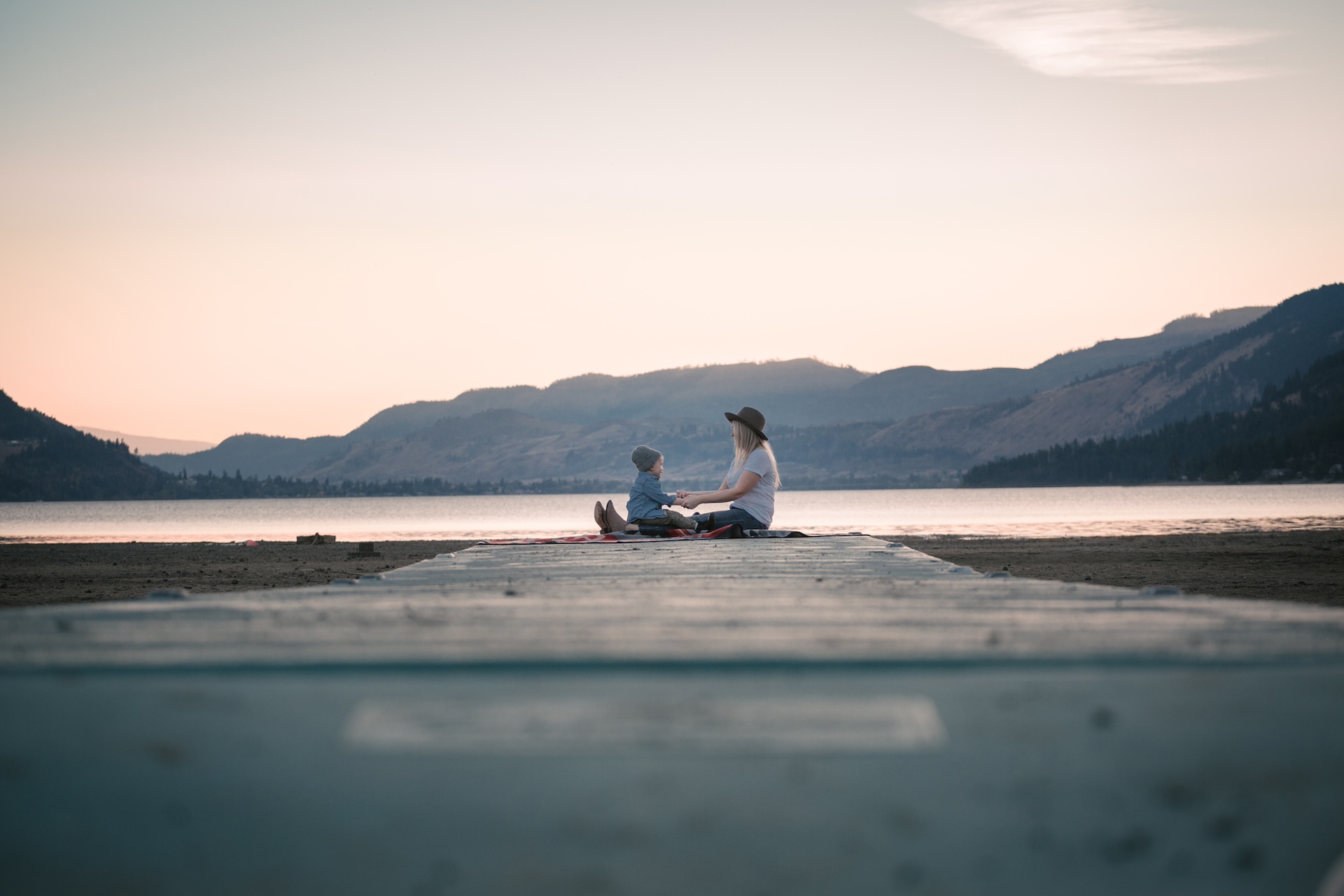A mother and child sit at the end of a boardwalk and hold hands next to the shore