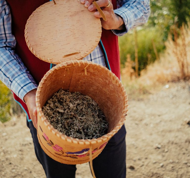 Person holding an open, woven basket filled with dried herbs or leaves.