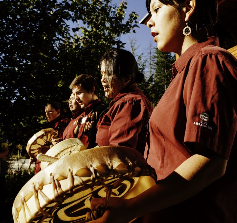 Individuals in maroon shirts holding decorated drums, outdoors with trees in the background.